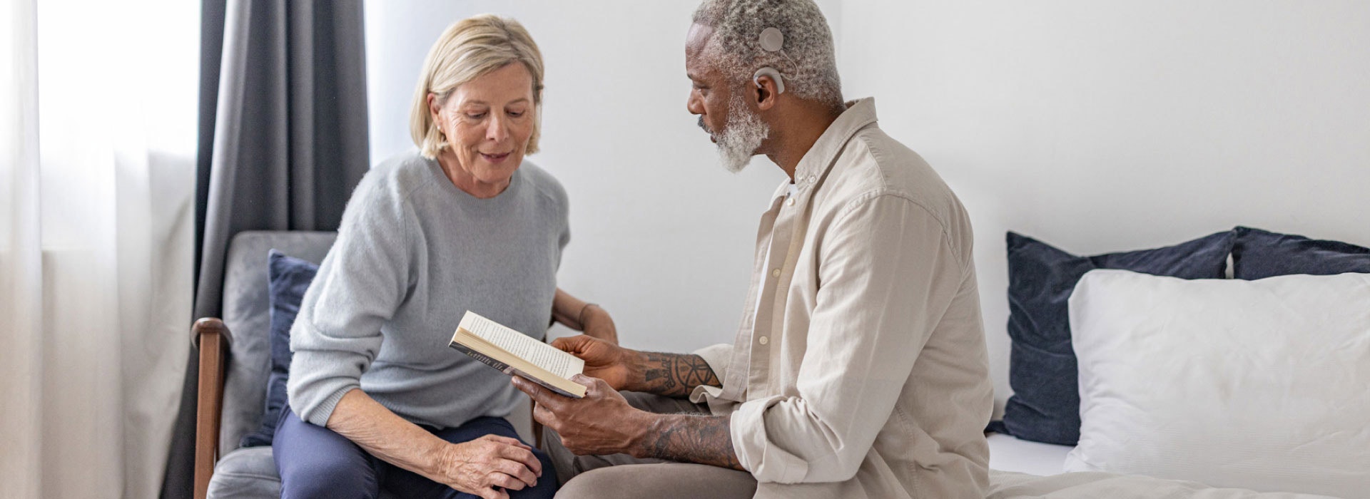 Elderly man reading to an elderly woman in a cozy room, illustrating the importance of hearing support like cochlear implants for single-sided deafness in later life.