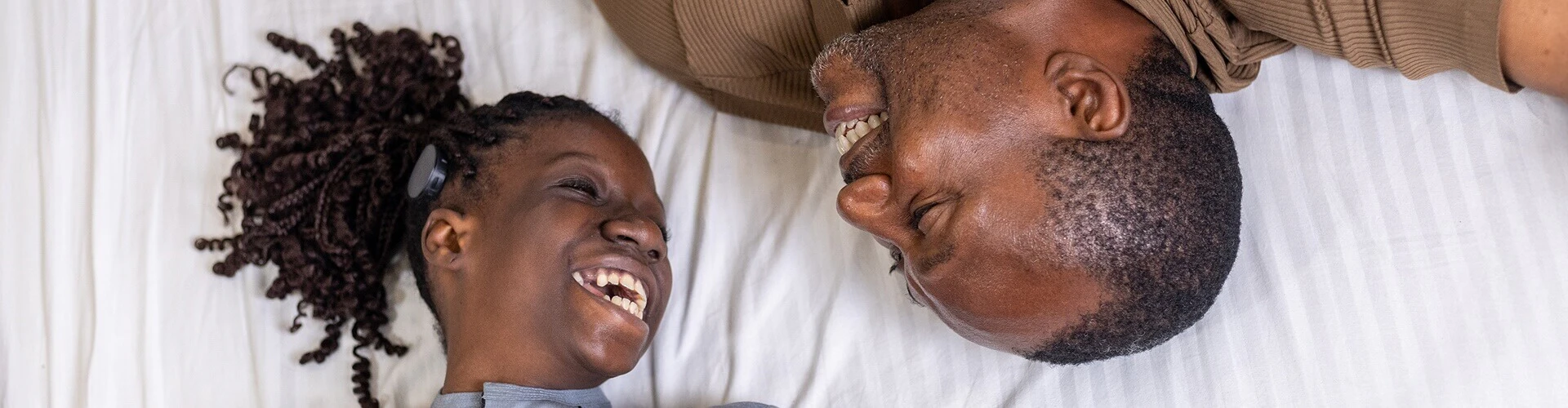 Two people laughing on a bed; left person wearing a RONDO 3 audio processor.