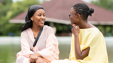 Two individuals sitting by the water and talking near a pavilion in a park.