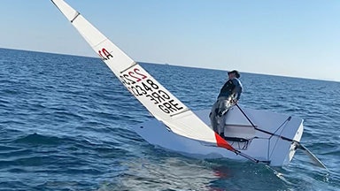 Ένα άτομο που πλέει σε ένα μικρό λευκό σκάφος με κεκλιμένο ιστό σε ανοιχτό νερό. Η θάλασσα είναι ήρεμη κάτω από έναν γαλάζιο ουρανό.