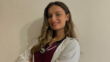 Μια γυναίκα που φοράει ένα λευκό εργαστηριακό παλτό πάνω από μια σκούρα κόκκινη κορυφή, με ένα στηθοσκόπιο τυλιγμένο γύρω από το λαιμό της, να στέκεται σε ένα απλό μπεζ τοίχο.