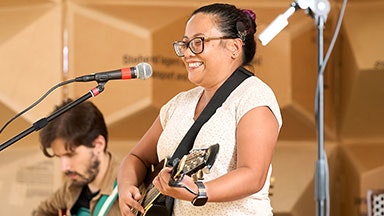 A woman with a cochlear implant playing an acoustic guitar and singing into a microphone on a stand. Another person with a guitar is partially visible in the background.