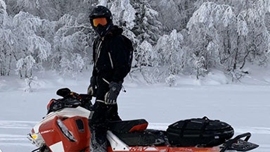 En person klædt i sort vinterudstyr og en hjelm, der står på en rød snescooter i et snedækket landskab med frostdækkede træer.
