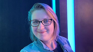 Een vrouw met kort blond haar die een lichtblauwe spijkerjas draagt, die voor een donkere achtergrond staat die door verticale neonblauwe lichten wordt verlicht.