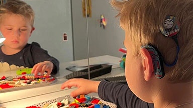 Et barn som leker med fargerike murstein på et bord, ser inn i et speil. En cochleaimplantatprosessor er synlig på øret hans.
