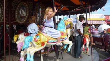 Un bambino in sella a un colorato cavallo di una giostra in un parco divertimenti. Altri bambini e adulti sono visibili nelle vicinanze, e la giostra ha decorazioni ornamentali.