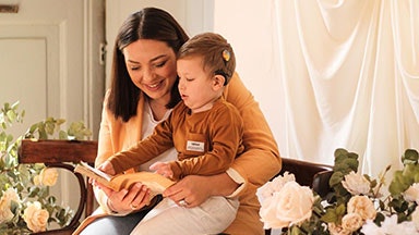 Un adulto seduto al chiuso mentre tiene un bambino in grembo leggendo un libro. Il bambino ha un audio processore per impianto cocleare sull'orecchio.