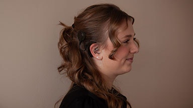 Een persoon met golvend bruin haar gestyled in een half-up look, het dragen van een zwarte top. Ze heeft een audioprocessor met een cochleair implantaat op haar oor.
