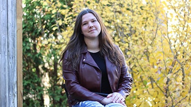 A person sitting outdoors on a wooden bench, wearing a brown leather jacket and blue jeans. The background features trees with yellow autumn leaves.