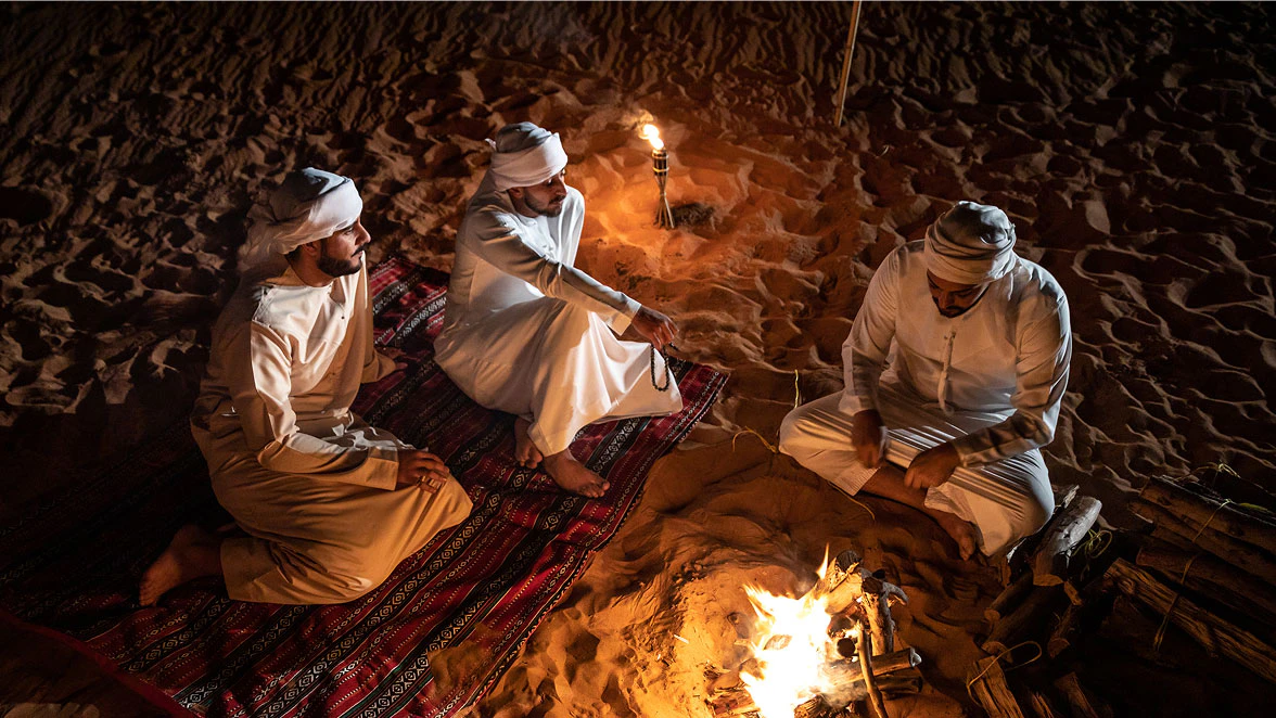 Three men sit around a campfire at night in the sand